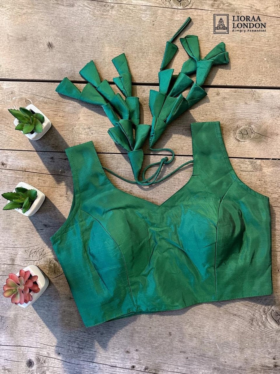 Lioraa London Ltd emerald green sleeveless silk blouse with back tie-up and decorative tassels on wooden background