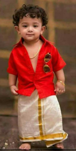Child wearing a red shirt and Velcro Onam Dothi traditional outfit with gold accents.